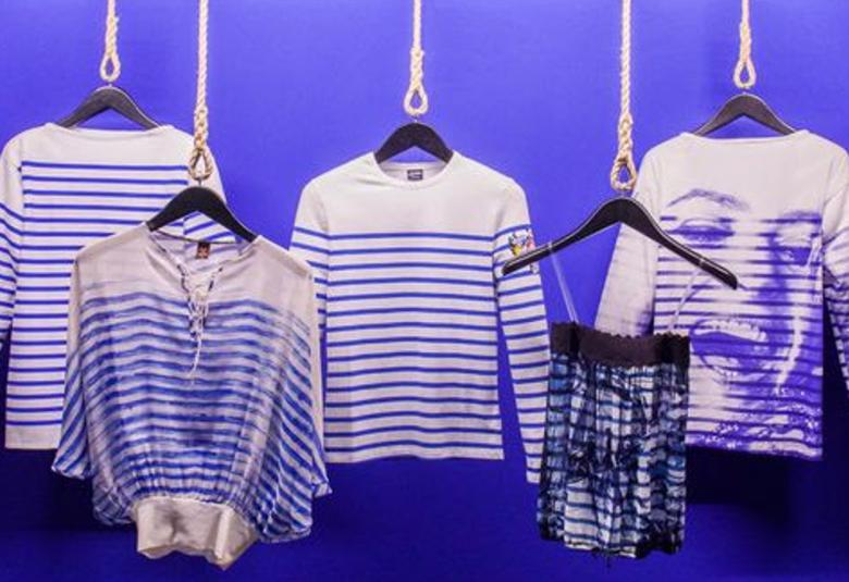 Kuvassa on kolme sini-valkoraitaista pitkähihaista paitaa riippumassa hengarissa. Yhdessä on nauravan naisen kasvot. Edessä vasemmalla on yksi sini-valkoraitainen leveä pusero ja vasemmalla raidallinen ja kuvallinen puolihame. Tausta on violetin sininen.