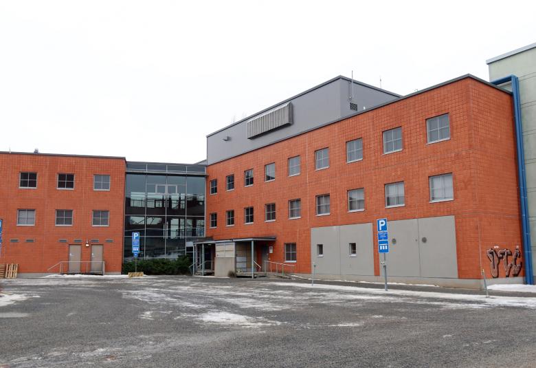 Kuvassa on punainen tiilinen kolmikerroksinen rakennus, joka on l-muotoinen.  Edessä on harmaata asfalttia.