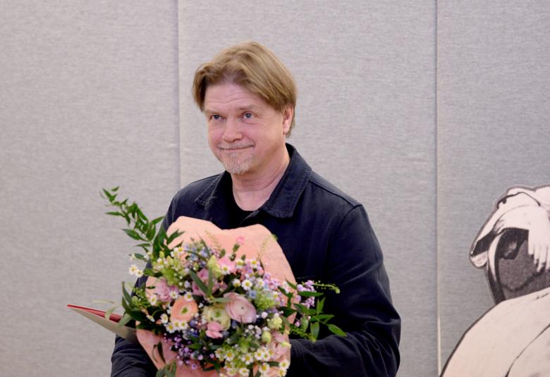 Kuvassa on Timo Parvela vaaleanpunainen kukkakimppu kädessään ja mustassa takissa.  Tausta on harmaa seinä.  Oikealla näkyy osittain isoa Minna Canthin piirroskuvaa.