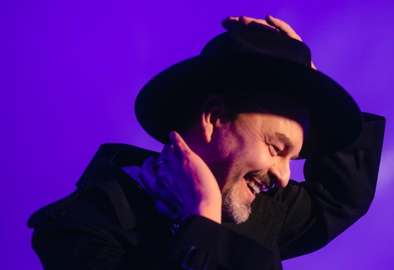 Kuvassa on Tuure Kilpeläinen sivukuvassa musta takki päällään ja musta lierihattu päässään. Hän pitää vasemmalla kädellään hatustaan kiinni. Tausta on violetti.