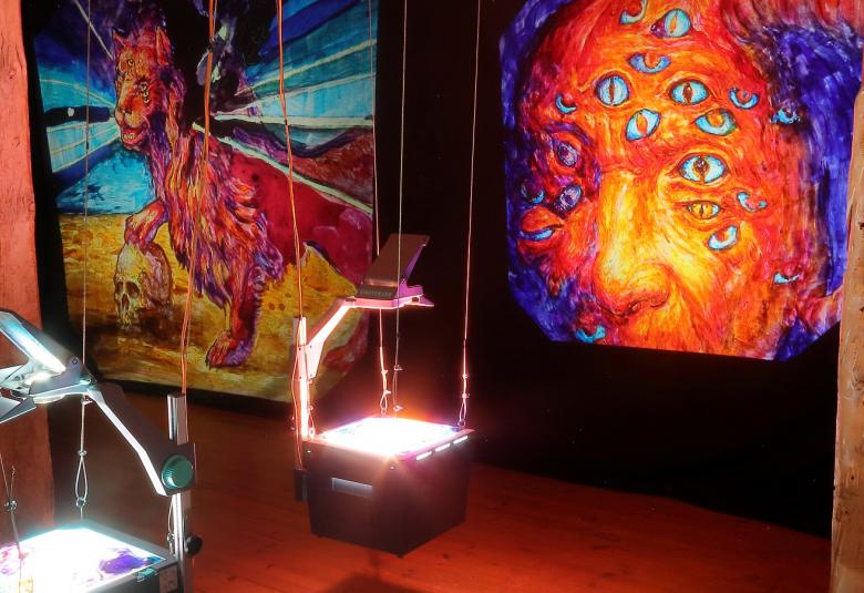 Kuvassa on taustalla seinällä oikealla isot, oranssin punaiset miehen kasvot, joissa on violetin värisiä silmiä ympäri kasvoja. Toisella seinällä  on oranssin punainen leijona, joka pitelee maassa olevaa pääkalloa oikealla tassullaan. Maa on keltainen ja leijonasta lähtee sinisiä säteitä. etualalla roikkuu kaksi piirtoheitintä.
