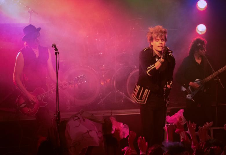 Kuvassa on bändi lavalla ja keskellä on Saku Taittonen laulamassa hieman kumartuneena eteenpäin. Molemmissa reunoiss aon kitaristit.  Lavalla on punertavaa savua ja valoa.  