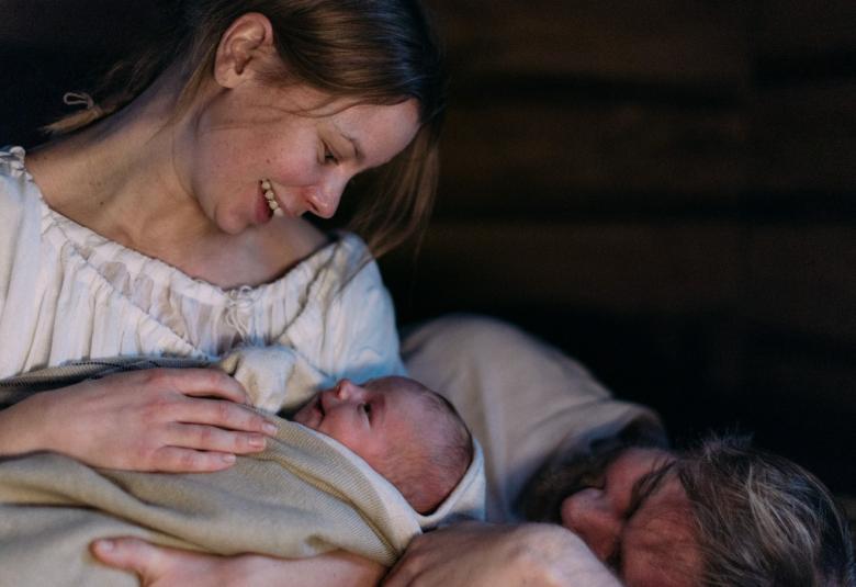 Kuvassa on Maija vauva sylissään ja hän katsoo sitä. Vieressä on sängyllä makaamassa pää tyynyllä on ja katsoo vauvaa. Tausta on tumma.