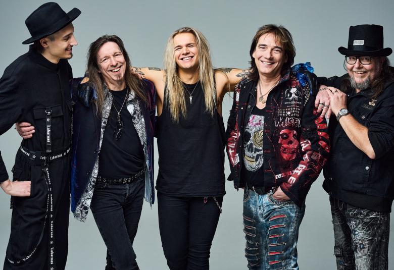 Kuvassa on Popedan viisi jäsentä seisomassa.  Heillä on mustat vaatteet tai kirjotut liivit tai pusakat päällään.  Vasemmalla olevalla miehellä on musta lierihattu päässään.