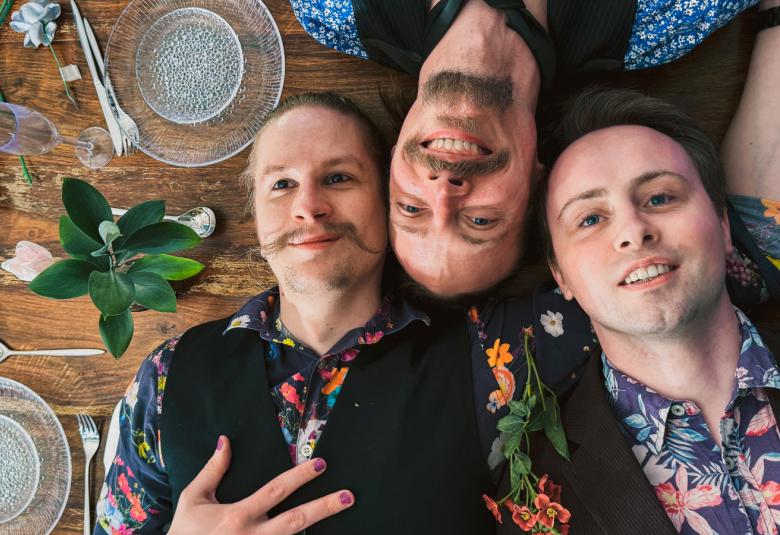 Kuvassa on kolme nuorta miestä, jotka makaavat asioilla kätetulla pöydällä. Kaksi miehistä on kasvot 'oikein' päin ja yksi nurinpäin. Heillä on kukalliset paidat ja mustat liivit.