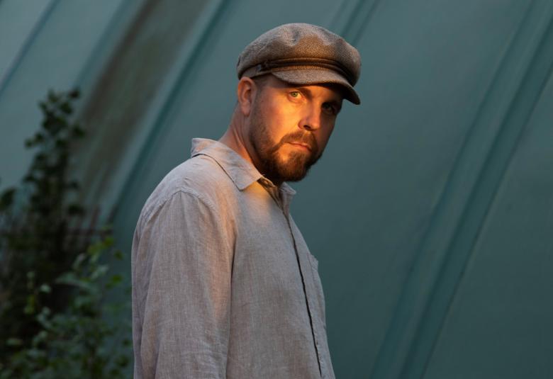 Kuvassa on Antti J. Leinonen ja hän on puolivartalokuvassa sivuttain ja katse kuvan katsojaan.  Hänellä on harmaa paita ja lippalakki päässään. 