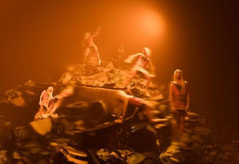 Kuvassa on roskakasan päällä yksi tanssija ja oikealla alempana kaksi tanssijaa.  Vasemmalla alempana vinottain on yksi tanssija kiven päällä. Kuvan sävy on ruskehtava.