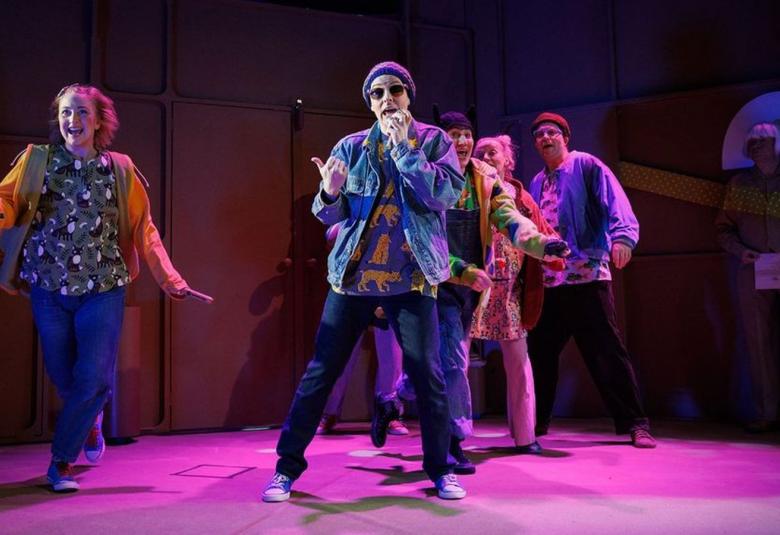 Kuvassa on vasemmalla tyttö, jossa on oranssi pusakka, vihreäkuvioinen paita ja siniset farkut.  Keskellä on on poika, jolla on sininen pipo ja farkkutakki ja tummat farkut. He tanssivat ja laulavat. Lattia on violetti. 