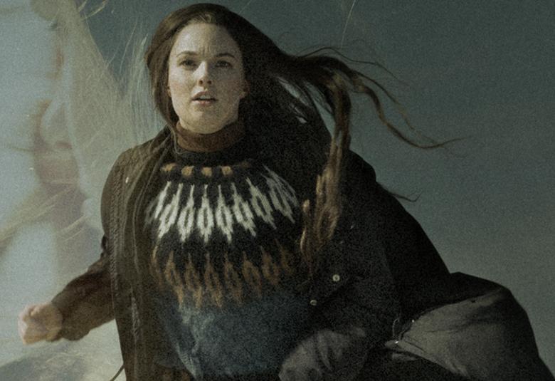 Kuvassa on Asta Sveholm liikkumassa sivuttain. Hänellä on ruskea norjalaisvillapaita, jossa on kuvioita. Ruskea takki on auki.  TAust aon ruskehtava ja Sveholmin pitkät hiukset liehuvat tuulessa.