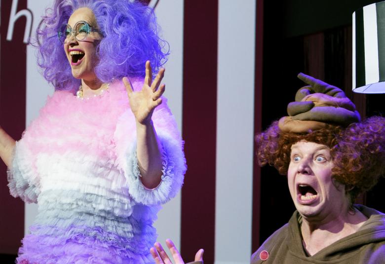 Kuvassa on vasemmalla nainen jolla on violetit kiharat hiukset ja pörhöinen vaalean punaisella, valkoisella ja violetilla raidoitettu mekko.  Oikealla istuu ruskeassa peikkomaisessa puvussa ja kädet levitettynä sekä päässään myssy.