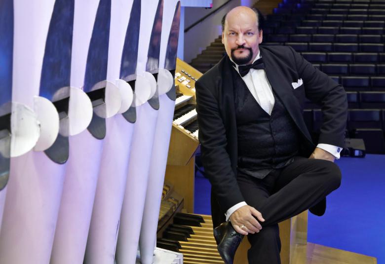 Kuvassa on vasemmalla vaalean violetin värisiä urkupillejä. Taaempana on Kalevi Kiviniemi istumassa urkujen äärellä ja kääntyneenä kuvan katsojaan päin. Hänellä on musta puku päällään. Taustalla näkyy tyhjää katsomoa.