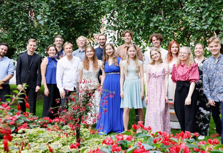 Kuvassa on seisomassa ryhmänä kesäisessä ympäristössä naisia ja miehiä kesävaatteissa. Taustalla näkyy vihreitä lehtipuita.