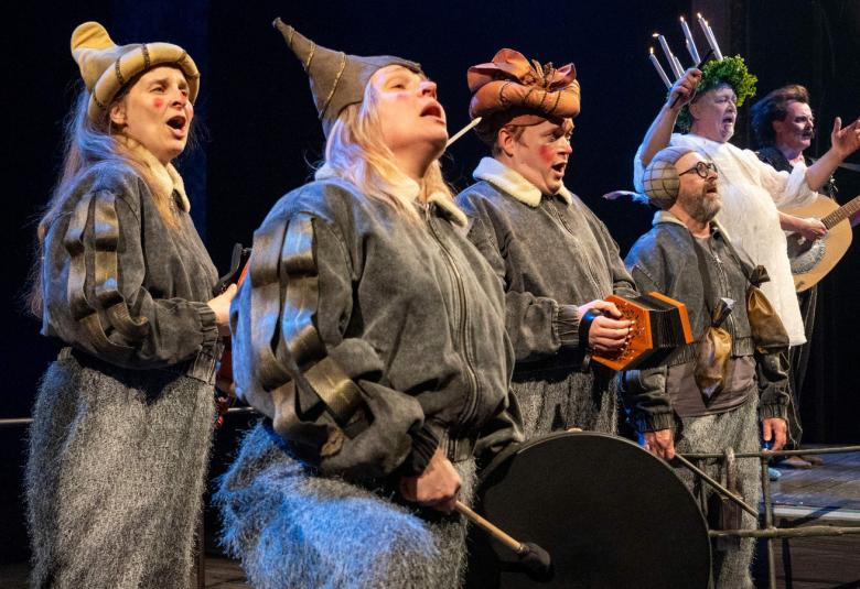 Kuvassa on harmaissa kokovartalon peittävissä, harmaissa puvuissa ja ruskehtavissa piippalakeissa neljä näyttelijää, joista etummainen soittaa rumpua. Taaempana on näyttelijä valkoisessa puvussa ja kynttelikkö päässään. Tausta on tumma.