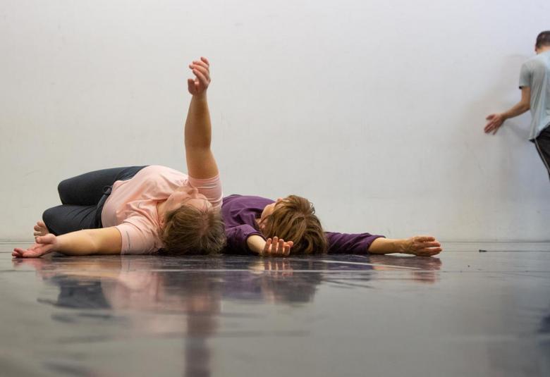 Kuvassa on kaksi naistanssijaa makaamassa lattialla ja vasemman puoleisella on oikea käsi ylhäällä. Taaempana vasemmalla on miestanssija nojaamassa harmaaseen seinään. Lattia on harmaan kiiltävä.