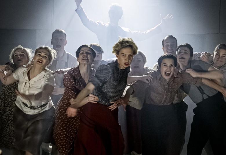 Kuvassa on tanssijoita kumartuneena eteenpäin vauhdikkaasti ja heillä on erivärisiä kauluspaitoja. Taustalla näkyy haamumaisesti yksi tanssija kädet ylhäällä ja hänen takaansa tulee hämyistä valoa. Kuvan sävy on tumma. 