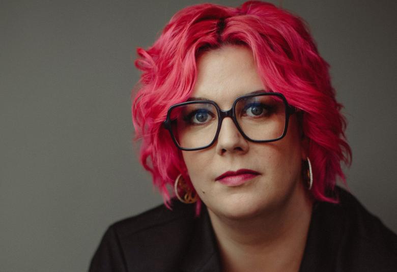 Kuvassa on Nilla Kjellsdotter kasvokuvassa ja hänellä on punaiset kiharaiset hiukset ja mustasankaiset isot silmälasit. Tausta on ruskehtavan harmaa.