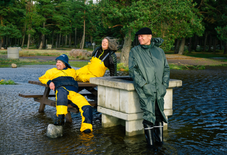 Kuvassa on vedessä keskellä neliön muotoinen vaalea grillipaikka ja sen vieressä pöytä penkeillä.  Vesi ylottuu pitkälle rantaan ja pöytä ja grillipaikka ovat myös vedessä. Penkillä istuu löhöten mies kelta-mustassa haalarissa ja tummassa sydvestissä. Pöydän päällä on keltaisessa vartalon yläosaan ulottuvassa haalarissa nainen ja grillipaikkaan nojaa mies tummassa sadetakissa mies. Taustalla on lehtipuita.