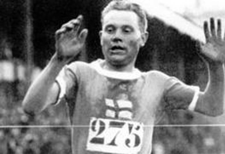 Kuvassa on Paavo Nurmi tulossa maaliin.  Hänellä on rinnassaan on numero 275. Hänen edessään on maalinauha.  Taustalla on himmeästi yleisöä.  Nurmella on kädet kohotettuna ylös.