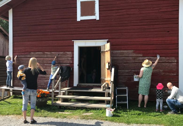 Kuvassa on punainen iso vanha varastorakennus, jossa on valkoiset ikkunan ja oven pielet sekä nurkkalaudat.  Sen edessä seisoo nainen vauvan kanssa ja alahirsiä on maalaamassa aikuisia ja lapsia.