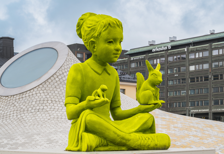 Kuvassa on vihreä veistos pikkutytöstä, joka istuu ja pitää vasemmalla kädessään iso oravaa ja oikeassa kädessään pientä oravaa.  Vasemmalla puolella on takana harmaa iso kartio, joss on pyöreä lasi vasemmalla. Oikealla näkyy taustalla ruskea tiilirakennus. 