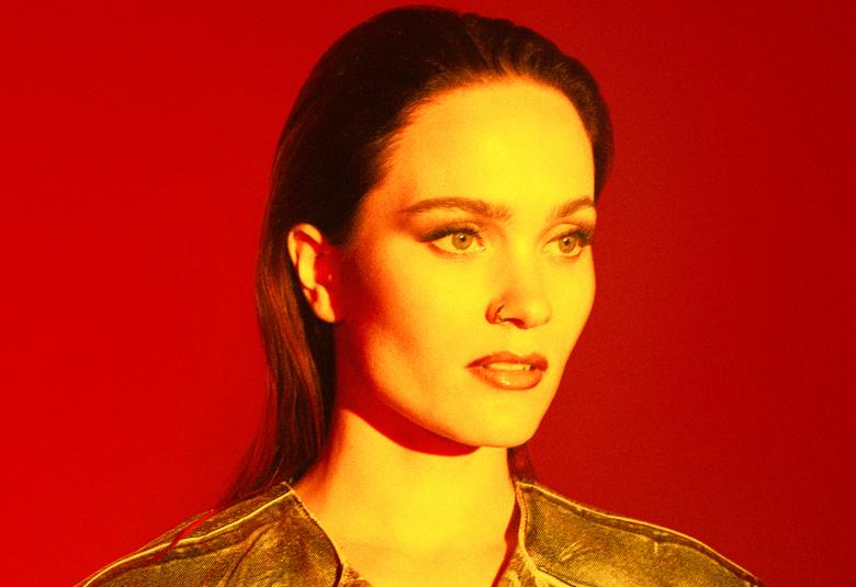 Kuvassa on Sanni kasvokuvassa. Hänellä on pitkät hiukset takana. Kuvan tausta on punainen kuvan sävy on kellertävä.