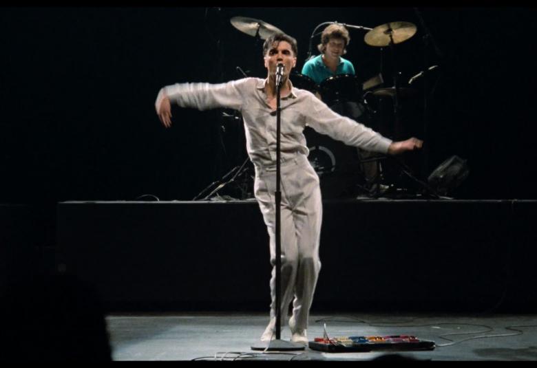 Kuvassa on mieslaulaja harmaassa paidass ja housuissa. Hän laulaa mikin takana ja tanssii kädet ojennettuina sivulle. Taustalla on tummaa. Takana on sinisissäKuvassa on vaatteissa oleva rumpali rumpusarjan takana.