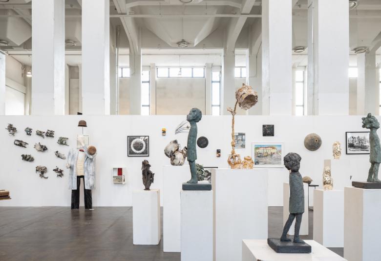 Kuvassa on korkea tila, jossa on taustalla valkoisia pylväitä.  Pylväiden luona on valkoisia taustoja ja niillä näkyy kehystettyjä valokuvia. Edessä keskellä on tummia veistoksia valkoisilla jalostoilla.