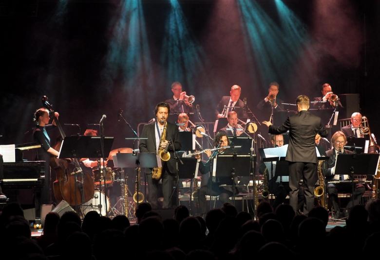 Kuvassa on UMO soittamassa yleisön edessä. Kuva on tumman sävyinen ja valaistuksen avulla näkyy orkesterin jäseniä. Etualalla näkyy mm. saksofonisti ja taustalla trumpetisteja.