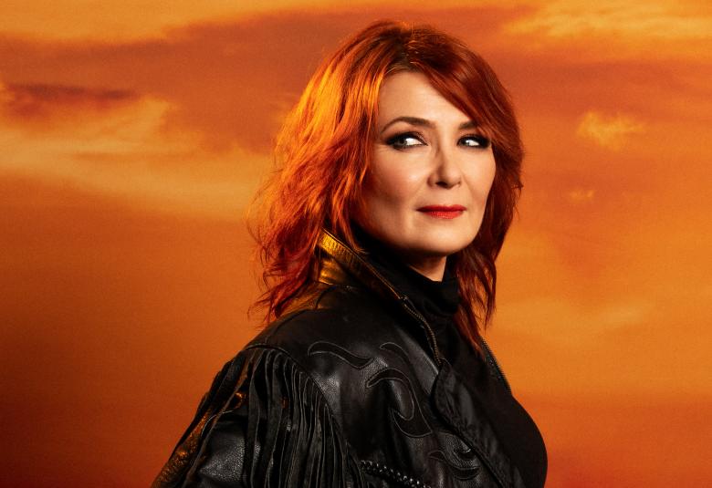 Kuvassa on keskellä on Irina puolivartalokuvassa mustassa nahkatakissa. Hänellä on punaiset hiukset ja hän katsoo vasemmalle.  Taustalla on oranssia taivasta. 