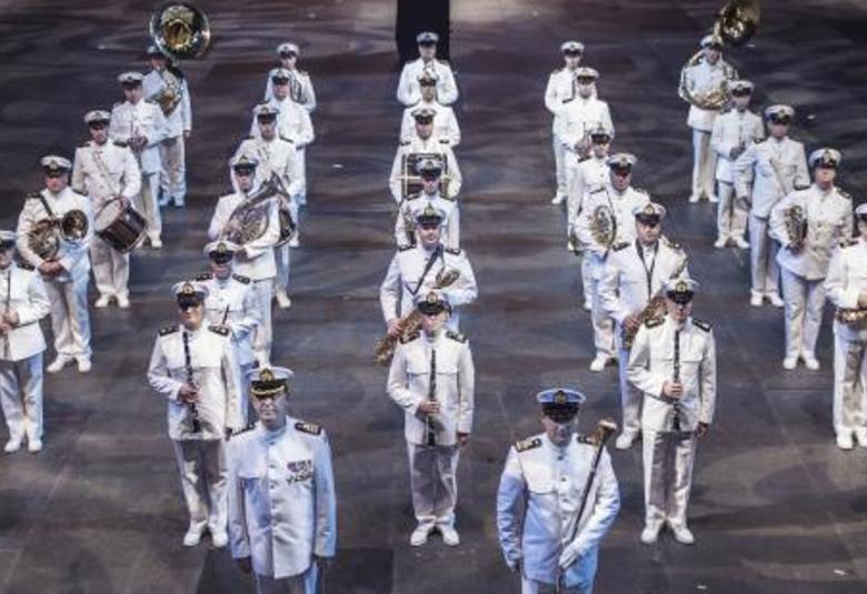 Kuvassa on Laivaston soittokunta valkoisissa puvuissa ja lakeissa seisomassa viidessä rivissä soittimet kädessään.  Kuva on otettu yläviistossa. Lattia on harmaa.