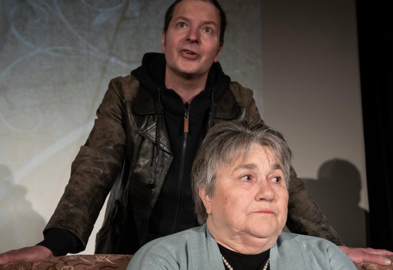 Kuvassa on takana mies violetissa takissa ja edessä nainen mustassa takissa kantamassa rullalla olevaa mattoa. Tausta on suurimmaksi osaksi tumma ja oikealla on vaaleaa.