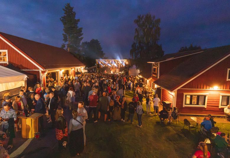 Kuvassa on maatalon pihapiiriä, jossa on punavalkoisia rakennuksia ja vasemmalla valkoinen teltta.  Pihapiirissä on paljon ihmisiä rakennusten välissä seisomassa ja taustalla on tumman sinistä iltataivasta loppukesällä.