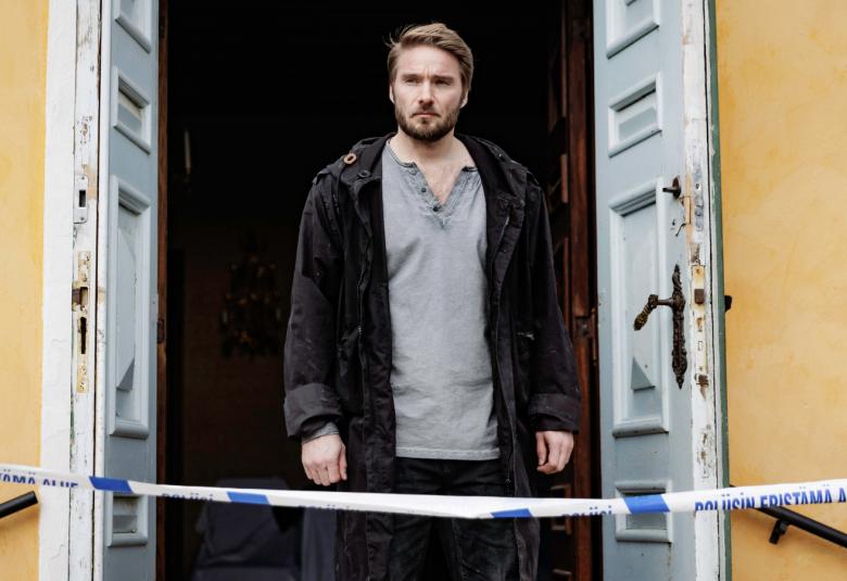 Kuvassa on Leo Sjöman seisomassa molemmin puolin avattujen vanhojen valkoisten ovien keskellä.  NIiden vieressä näkyy keltaista seinää. Edessä alempana on sini-valkoista poliisinauhaa.