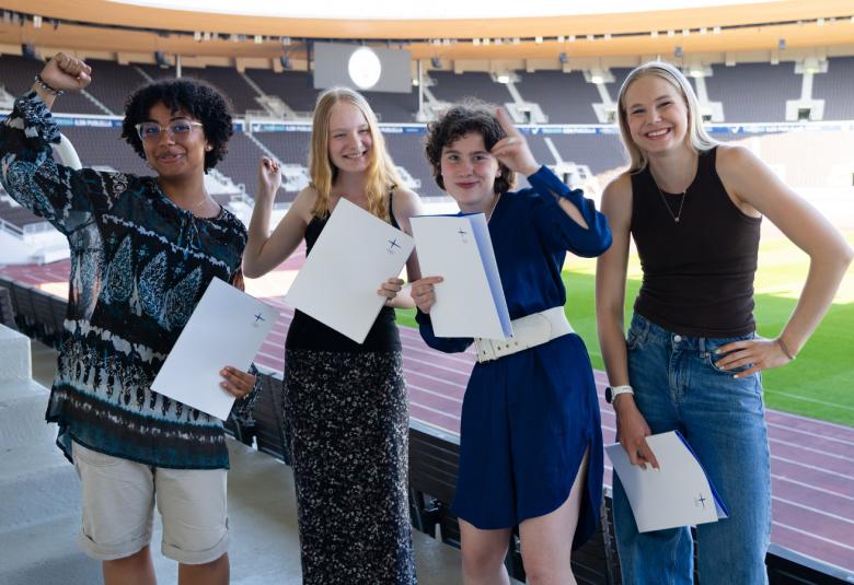 Kuvassa on neljä tyttöä kunniakirjat kädessään. Vasemmalla on tumma ihoinen tyttö ja hänellä on sinivalkoinen pusero ja valkoiset shortsit. Vieressä olevalla on pitka, sininen mekko ja seuraavalla tumman sininen ja reunummaisellä musta hihaton t-paita sekä siniset farkut.