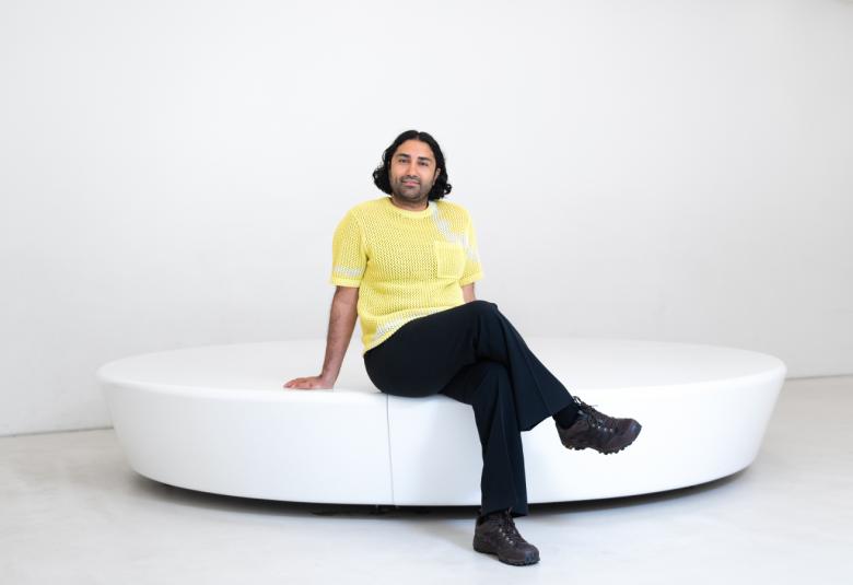  Kuvassa on Harold Hejazi, joka istuu valkoisen ison soikean istuimen päällä. Hänellä on keltainen t-paita ja mustat housut. Seinä ja lattia ovat valkoisia.