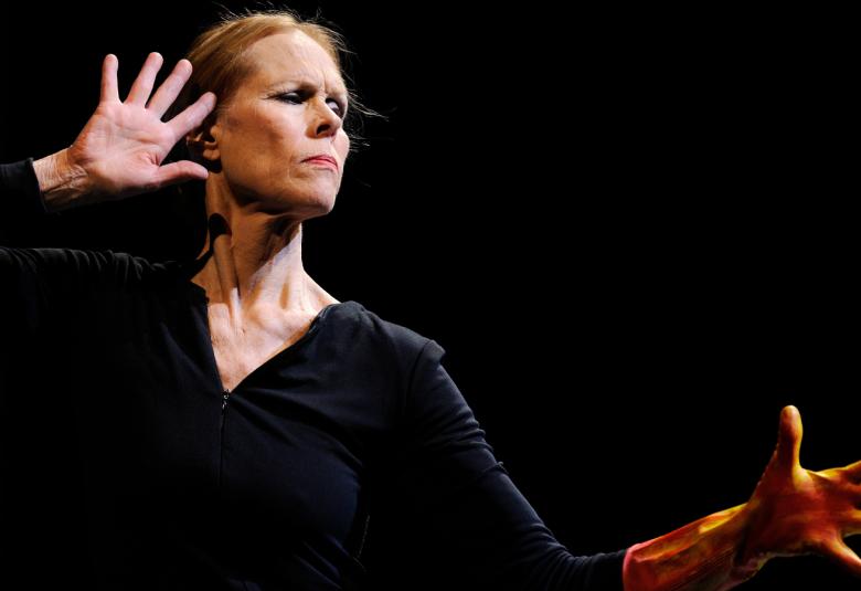 Kuvassa on Carolyn Carlson puolivartalokuvassa tanssimassa ja hänellä on musta pusero. Hänellä on oikea käsi harallaan olevin sormin pään kohdalla.  Tausta on musta.