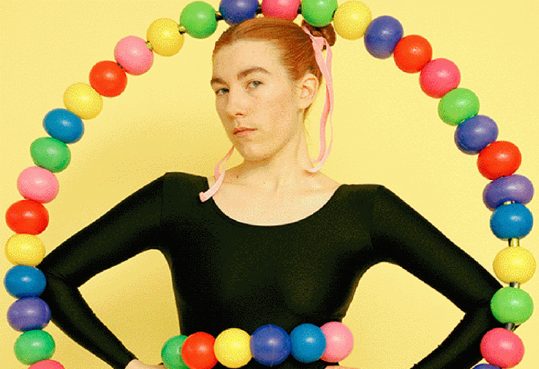 Kuvassa on Emma Sarpaniemi mustassa tiukassa paidassa puolivartalokuvassa. Hänen ympärillään menee värikkäistä palloista tehty rengas ja hänen vyötäröllään on myös pallorengas. Tausta on keltainen.