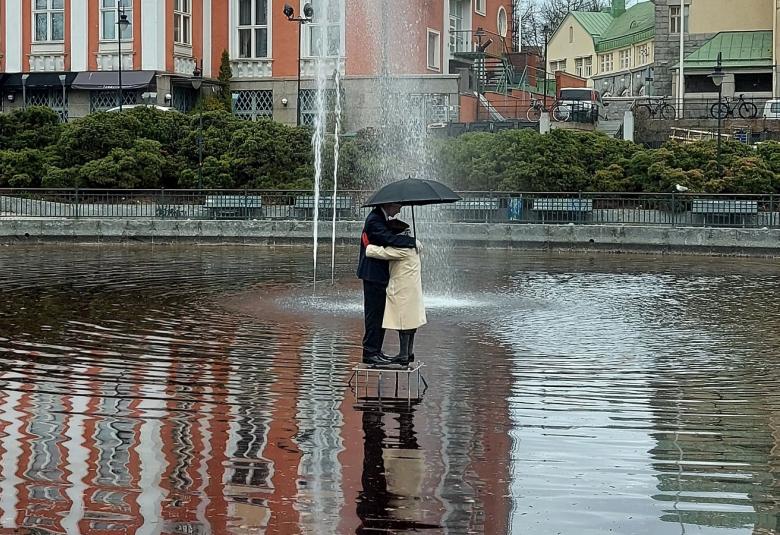 Kuvassa on suihkulähde ja sen ympärillä laajasti vettä.  Keskellä on halaava pariskunta sateenvarjon alla.  Taustalla on ylempänä oranssi ruutuikkunainen, suuri talo.