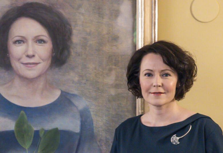 Kuvassa on vasemmalla Jenni Haukion muotokuvaa ja hänellä on siinä sini-harmaata taivasta kuvaava pusero, jossa on alempana järvä ja kaksi vihreätä lehteä.  Hän seisoo alempana itse ja hänestä on puolivartalokuvaa.  Hänellä on tumman sininen mekko päällään ja siinä on hopeinen rintakoru.