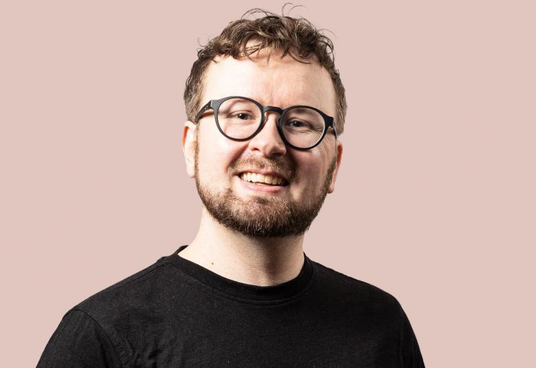 Kuvassa on Jukka Aalto puolivartalokuvassa ja hänellä on musta t-paita päällään sekä musta sankaiset silmälasit. Tausta on vaalean ruskehtava.