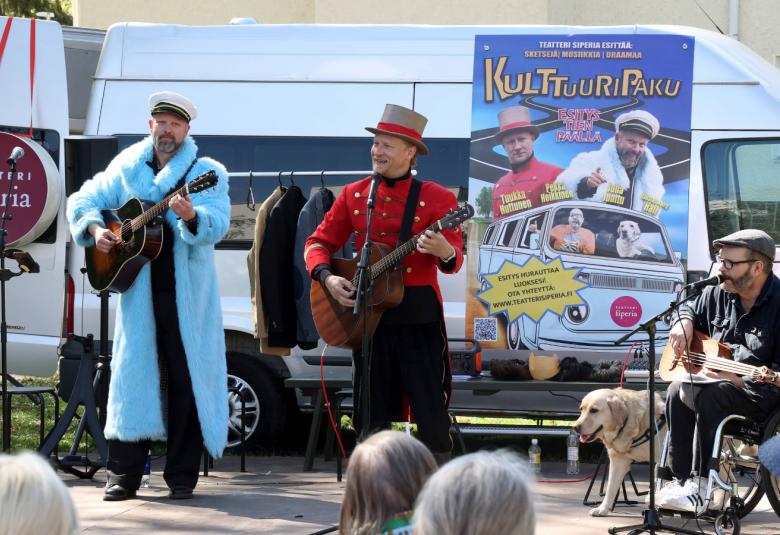 Kuvassa on valkoisen pakun edessä kolme miesesiintyjää. Vasemman puoleisella on valkoinen pitkä takki ja hän soittaa kitaraa.  Keskellä on punaisessa puserossa ja siliterissä mies, joka soittaa myös kitaraa. Oikealla istuu mies soittamassa kitaraa tummassa puvussa vierellään valkoinen koira. Taustalla näkyy pukun seinällä mainosjuhliste.