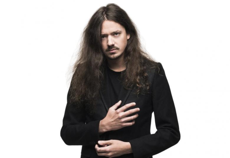 Kuvassa on Lauri Porra puolivartalokuvassa mustassa takissa. Hänellä on pitkät kiharat hiukset. Kuvan tausta on valkoinen.