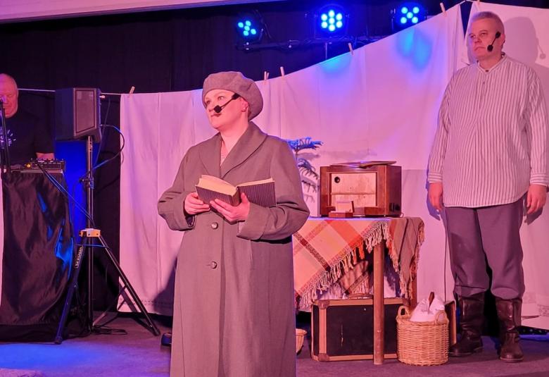 Kuvassa on taustalla pyykkinarulla roikkuu lakanoita. Edessä on pöytä, jonka päällä on liina ja vanha radio. Pöydän vierässä on mies seisomassa vaaleassa paidassa.  Edessä on nainen harmaassa takissa ja baskerissa seisomassa sekä kirja kädessään.  Taustalla on seisoo vasemmalla mies sinisessä paidassa. Kuva on punasävytteinen.