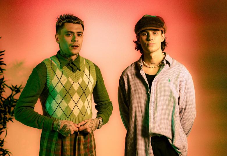 Kuvassa on vasemmalla Mikael Gabriel harmaa-valkoruudullisessa liivissä ja harmaassa paidassa. Oikealla jambo vaalean ruskeassa paidassa ja tummassa lippalakissa. Molemmat seisovat puolivartalokuvassa. Tausta on oranssi.