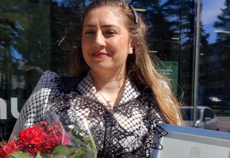 Kuvassa on Natasa Autio ja hänellä on pitkät tummat hiukset sekä päällään musta-valkoinen pikkuruudullinen pitkähihainen pusero. Hänellä on oikeassa kedessään tumman punaisia ruusuja ja vasemmassa kunniakirja. 