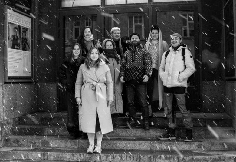 Kuvassa ovat KOM-teatterin portailla syvennyksessä seisomassa ulkovaatteissa. Vasemmalla on seinällä vaalea juliste ja seinään on kiinnitetty pystysuorassa teatterin nimi KOM teatteri. Edessä näkyy lumisadetta.