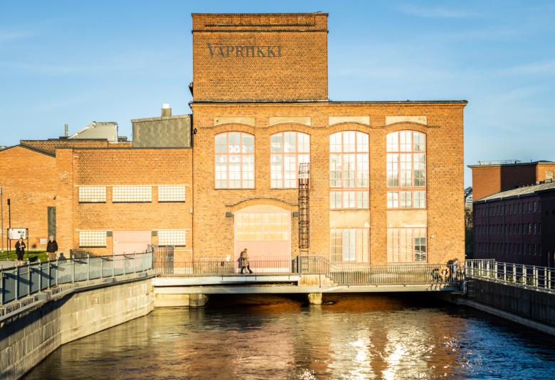 Kuvassa on Tampereen Vapriikin tehdasrakennus ja se on ruskeatiilinen.  Edessä on vesipato. Taustalla on sininen taivas.