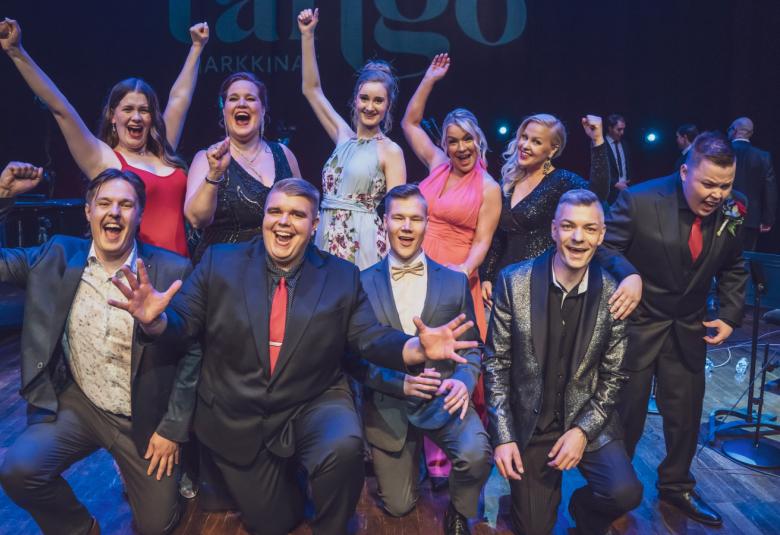 Kuvassa ovat Tantomarkkinoiden finalistit yhtieskuvassa. Siinä ovat naisfinalistit seisomassa takana ja lähes kaikki viisi tuulettavat toinen käsi ylhäällä. Edessä ovat miehet istumassa iloisina. Kuvassa on tumman sinistä valaistusta.