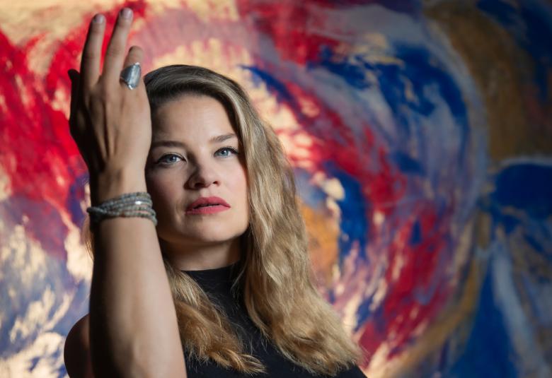 Kuvassa on vasemmalla Marjukka Tepponen puolivartalokuvassa ja hänellä on oikea käsi pystyssä.  Tepposella on vaaleahkot pitkät hiukset. Taustalla on värikkään kirjava seinämää.