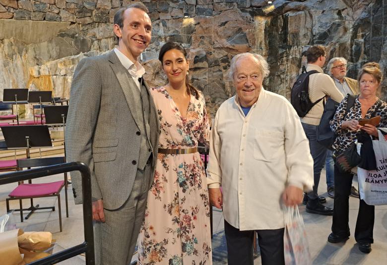 Kuvassa ovat Luck Terence harmaassa puvussa, Iris Candeleria vaalean roosan värisessä kukallsessa mekossa ja Jorma Panula valkoisessa paidassa. Taustalla kallioseinämää ja muita ihmisiä.